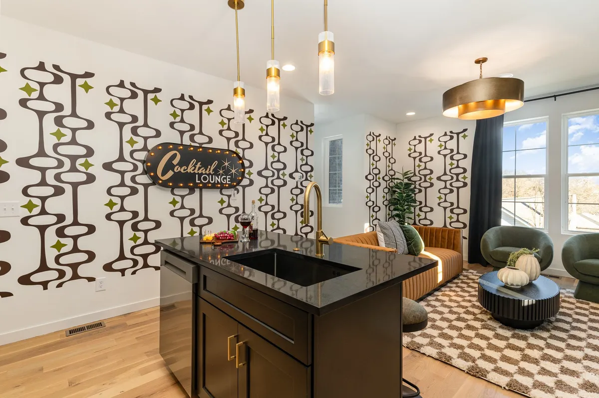 The image showcases a stylish interior lounge area featuring a playful 'cocktail lounge' neon sign, a sleek black kitchen island perfect for mixing drinks, plush green accent chairs for relaxing, and unique decorative wall patterns—all arranged to encourage socializing and entertaining.