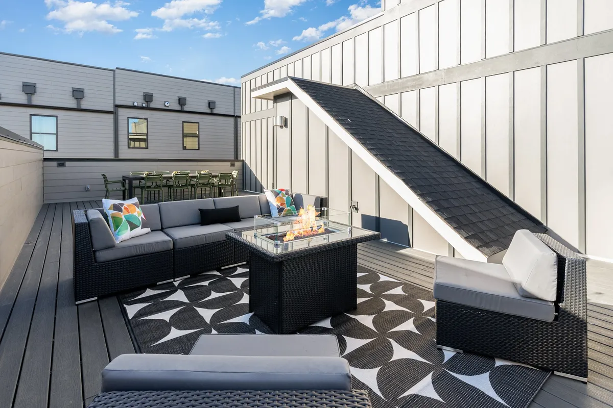 This image features a contemporary rooftop space with a fire pit as the focal point, surrounded by stylish outdoor seating and a decorative rug. A staircase and open sky frame the setting, suggesting a welcoming place for evening gatherings and stargazing.