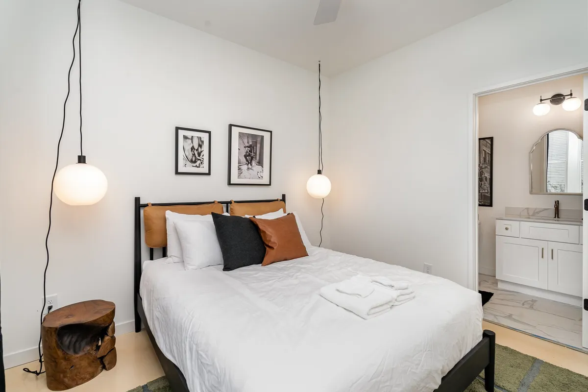 A serene bedroom featuring a stylish bed with hanging lights, tasteful artwork, and a natural wooden stool. Morning sunlight softly illuminates the space, creating a calm and restful atmosphere ideal for relaxation.