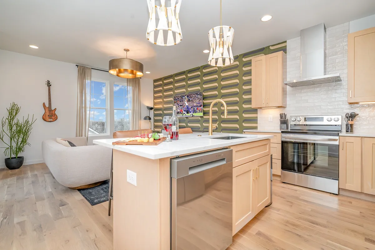 This image features a kitchen island with bar seating, unique wallpaper, contemporary lighting, a guitar as decor, and large windows that flood the space with natural light. The adjacent living area offers a comfortable spot for relaxing or socializing while watching TV.